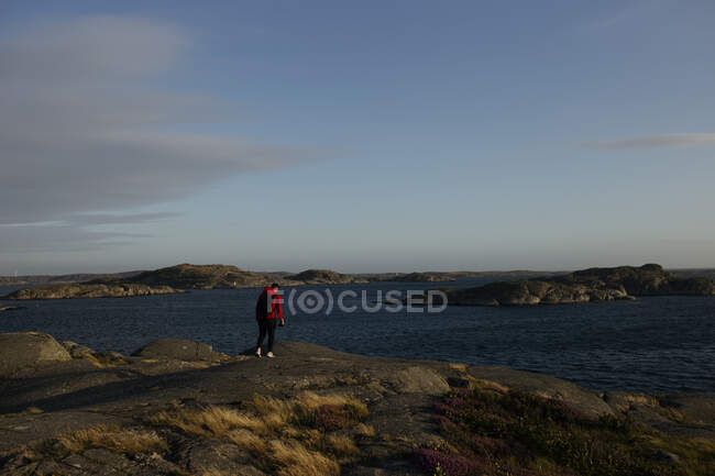 Back view of unrecognizable male traveler with backpack in casual wear standing with photo camera on rocky surface near lake — Stock Photo