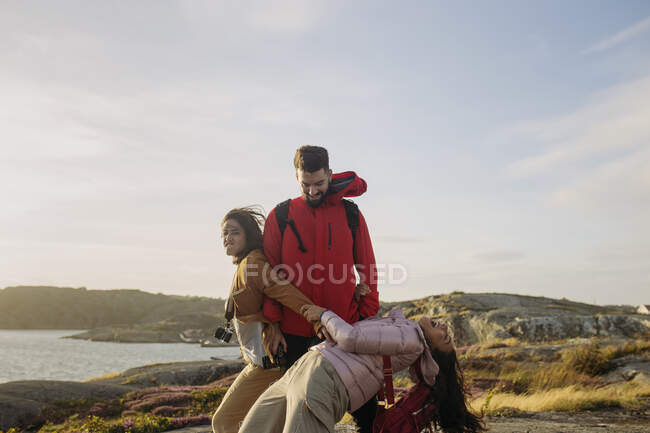 Grupo de jovens turistas alegres em outerwear casual e com mochilas e câmera de fotos de pé juntos e de mãos dadas em penhasco rochoso na praia ensolarada — Fotografia de Stock