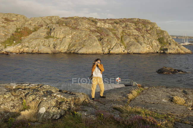 Anonimo giovane viaggiatore femminile in abiti casual in piedi sulla scogliera rocciosa sulla riva del mare e scattare foto di paesaggi incredibili durante il viaggio — Foto stock