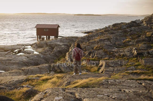 Back view of anonymous female tourist in casual outerwear and with backpack standing alone on rocky cliff on seashore and admiring amazing landscape — Stock Photo