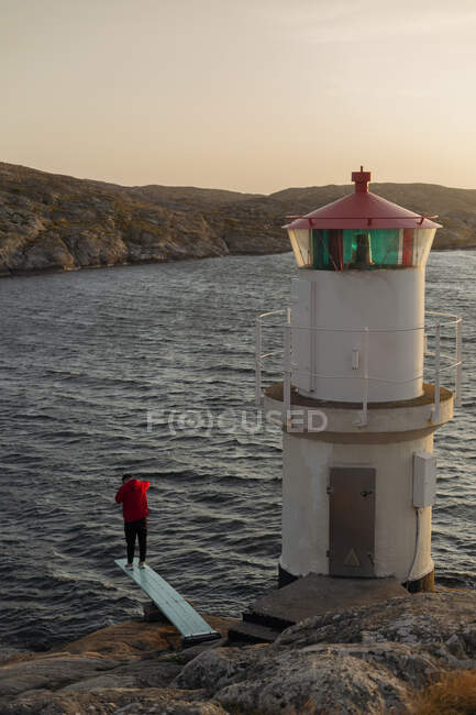 Back view of anonymous male tourist in red jacket standing alone on edge of wooden pier and looking down at water near lighthouse on rocky seashore — Stock Photo
