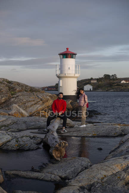 Adult man in red jacket sitting on fallen tree trunk and woman in casual clothes standing on stones near river and lookout tower looking at camera on cloudy day — Stock Photo
