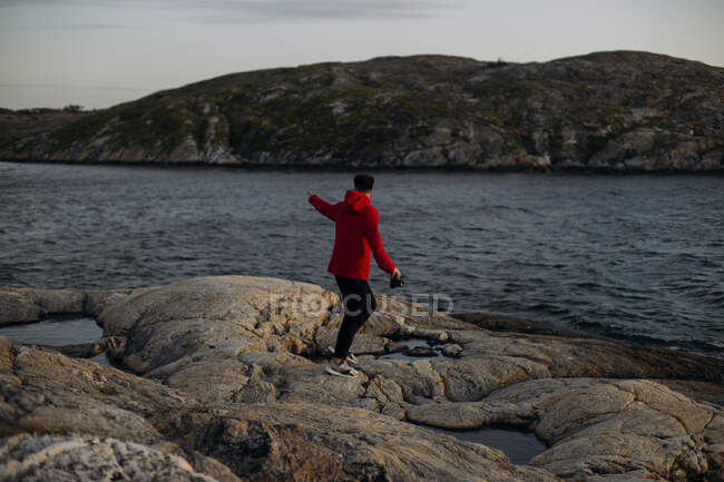 Вид збоку невпізнаваного чоловіка-мандрівника в повсякденному одязі, що стоїть з фотоапаратом на кам'янистій поверхні біля озера та вежі спостереження — стокове фото