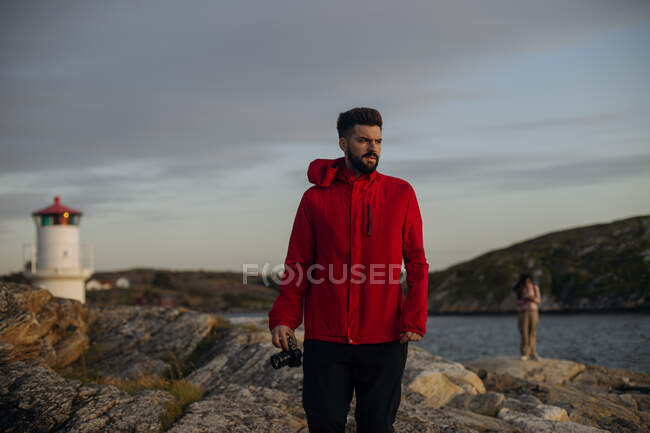 Серйозний чоловік-мандрівник у повсякденному вбранні стоїть з фотоапаратом на кам'янистій поверхні біля озера та вежі спостереження і дивиться вбік — стокове фото