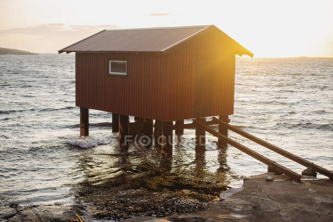 Коричневая плавучая лодка с небольшим окном и дверью, установленная на понтоне недалеко от морского побережья, с волнами, касающимися поверхности берега — стоковое фото