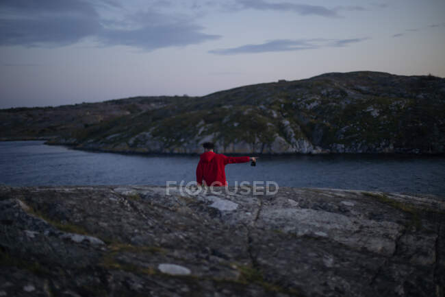 Visão traseira do turista remoto com capuz casual vermelho em pé em penhasco rochoso perto do mar e apontando o dedo para o lado, segurando a câmera na mão — Fotografia de Stock