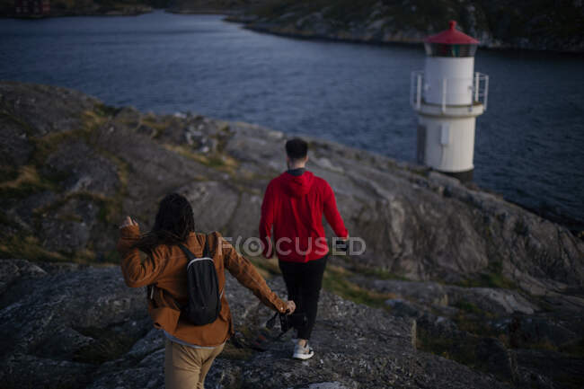 Обратный вид анонимной пары путешественников в повседневной одежде с фотокамерами в руках, идущих по скалистому побережью к морю и любующихся удивительным видом — стоковое фото