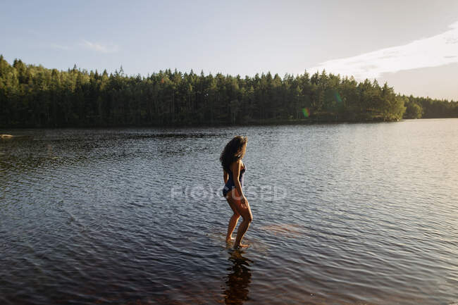 Вигляд стрункої самиці в купальнику стоїть у спокійній воді озера, насолоджуючись заходом сонця і величними краєвидами. — стокове фото