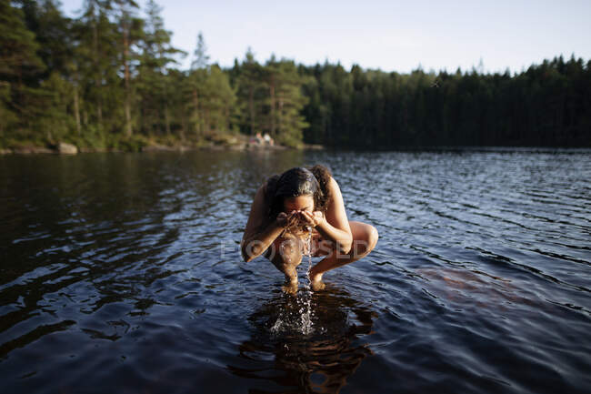 Unrecognizable female squatting barefoot in water and washing face on background of majestic forest during sunset — Stock Photo