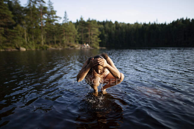 Unrecognizable female squatting barefoot in water and washing face on background of majestic forest during sunset — Stock Photo