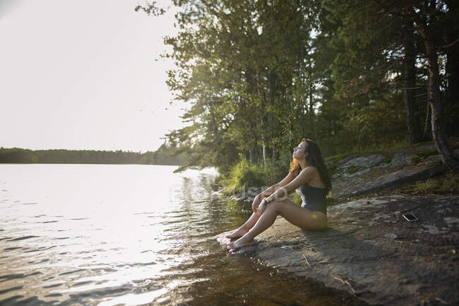 Vue latérale de la femelle aux yeux fermés portant un maillot de bain assis sur la rive rocheuse du lac et profitant d'un paysage pittoresque de forêt — Photo de stock