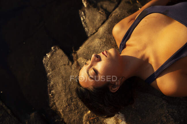 Von oben eine entspannte Frau im nassen Badeanzug, die mit geschlossenen Augen am felsigen Seeufer liegt und im Sommerurlaub den Sonnenuntergang genießt — Stockfoto