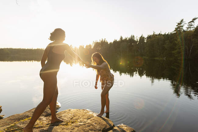 Mujeres jóvenes en traje de baño cogidas de la mano después de nadar en el lago durante las vacaciones de verano - foto de stock