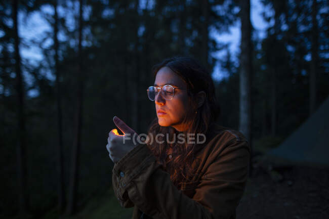 Serena campeggiatrice indossa giacca e occhiali in piedi in legno e guardando la fiamma da accendino al buio — Foto stock