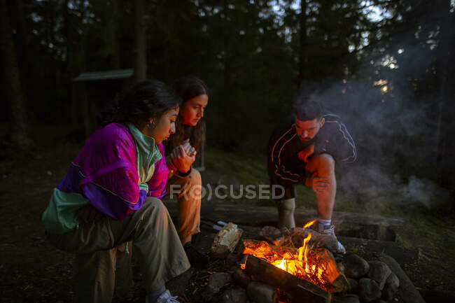 Company of friendly people in casual clothes gathering around bonfire in wood while making fire and warming up in evening — Stock Photo