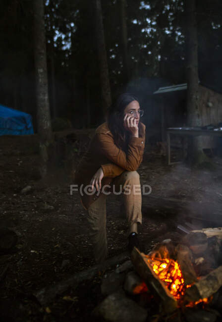 Camper femenina en traje casual de pie cerca de la fogata y relajarse durante el camping mientras reflexiona y mirando hacia otro lado - foto de stock
