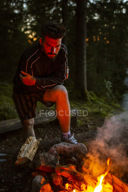 Tranquillo camper maschile in abbigliamento casual in piedi con log vicino al falò di notte e riscaldamento durante il campeggio in legno — Foto stock