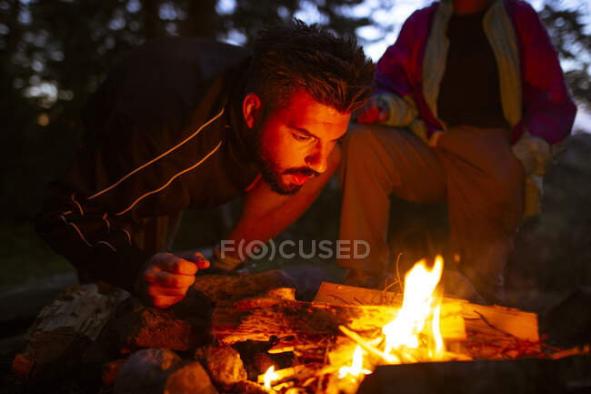 Спокойный мужчина-кемпер в повседневной одежде стоит с бревном возле костра ночью и согревается во время кемпинга в лесу — стоковое фото