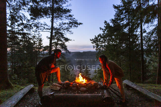 Pareja joven acampando juntos en ropa casual con tronco cerca de la hoguera al atardecer y calentándose durante el campamento en madera - foto de stock
