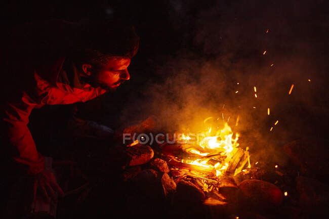 Ruhiger männlicher Camper in Freizeitkleidung, der nachts mit Baumstamm am Lagerfeuer steht und sich beim Zelten im Wald aufwärmt — Stockfoto