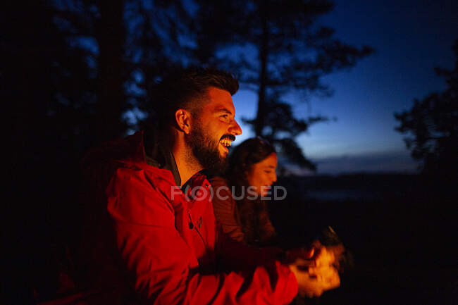 Jeune couple campant ensemble en tenue décontractée avec bûche près du feu au coucher du soleil et se réchauffant pendant le camping dans le bois — Photo de stock