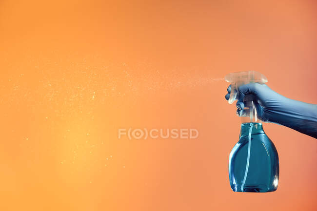Persona anónima con spray en botella de plástico para desinfección de superficies pulverizando líquido sobre fondo naranja en estudio - foto de stock