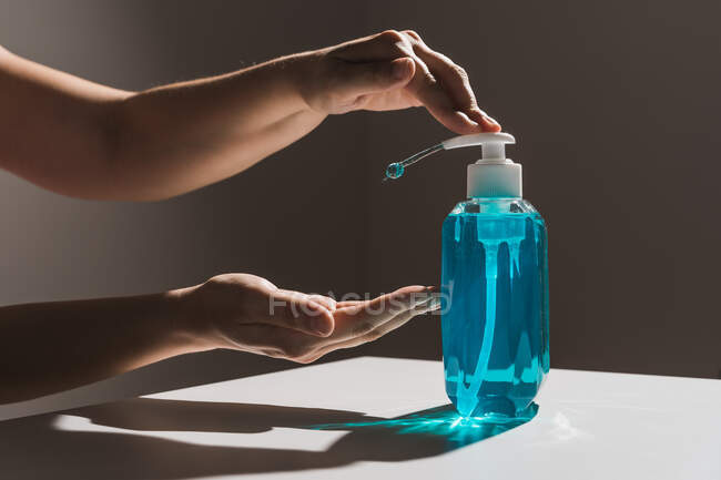 Crop personne sans visage tirant les mains pour bouteille de savon liquide en plastique bleu avec distributeur blanc à l'intérieur situé sur la table blanche avec ombre — Photo de stock