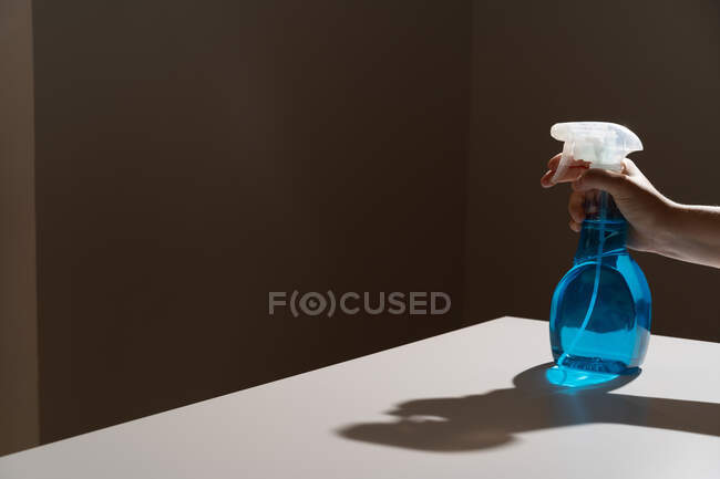 Crop anonymous person pushing dispenser mechanism and directing white cleaner flow on black wall while bottle being placed on white table with shadows — Stock Photo
