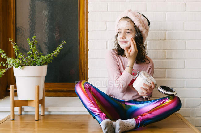 Позитивная маленькая девочка в повседневной одежде и розовой повязке сидит на столе с горшком и наносит маску для лица из стеклянной банки на фоне белой кирпичной стены с деревянным окном — стоковое фото