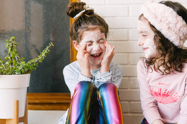 Joyful laughing girls in similar clothes and applied face mask sitting on wooden table at home having fun against brick white wall — Stock Photo