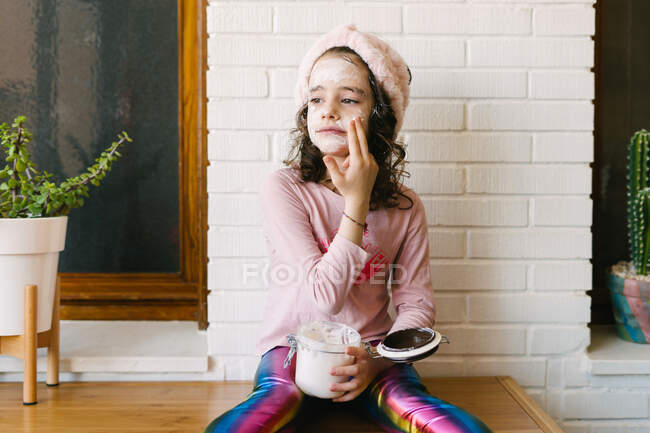Позитивна маленька дівчинка в повсякденному одязі і рожева пов'язка, що сидить на столі з горщиком для будинку і наносить маску для обличчя зі скляної банки на тлі білої цегляної стіни з дерев'яним вікном — стокове фото