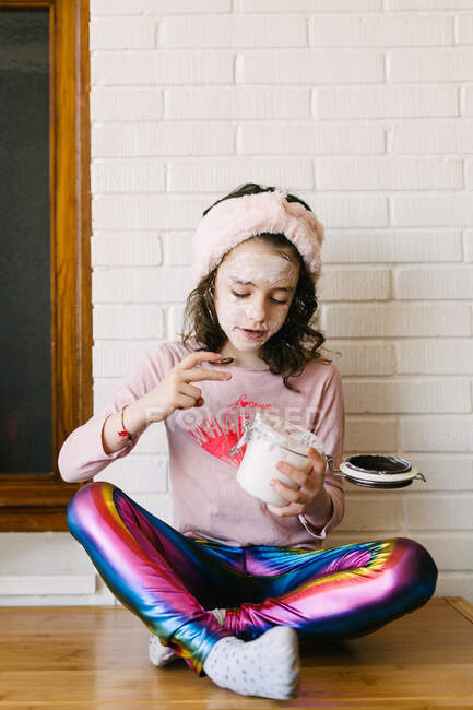 Positive little girl in casual clothes and pink headband sitting on table with potted houseplant and applying face mask from glass jar against background of white brick wall with wooden window — Stock Photo