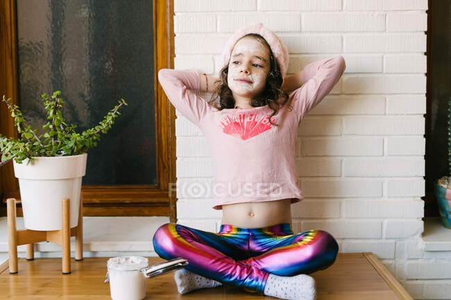 Счастливая улыбающаяся маленькая девочка с вьющимися волосами в розовой пижаме и повязкой на голове, сидящей возле белой кирпичной стены в маске для лица — стоковое фото