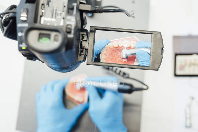 Desde arriba del aprendiz de cultivo en guantes azules realizando operación dental con brote y utilizando microscopio de operación médica con cámara digital - foto de stock