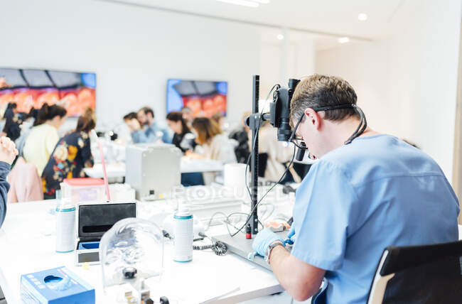 Rückansicht eines männlichen Zahnmedizinstudenten in Uniform und Handschuhen, der mit dem Mikroskop arbeitet, während er im modernen Klassenzimmer zahnärztliche Operationen durchführt — Stockfoto