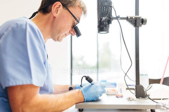 Vista lateral del estudiante de odontología masculino en uniforme y guantes trabajando con microscopio mientras realiza la operación dental en el aula moderna - foto de stock