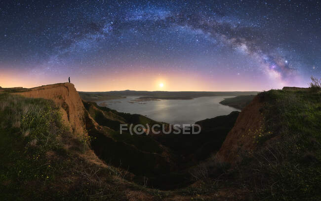 Erstaunliche Schlucht mit Silhouette einer unkenntlichen Person, die in einer Klippe in der Nähe des Sees unter buntem Nachthimmel mit Milchstraße auf dem Hintergrund steht — Stockfoto