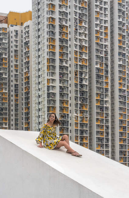 Mulher relaxada em vestido multicolorido sentado no telhado inclinado concreto e olhando para a câmera com arranha-céus residenciais no fundo em Hong Kong, na China — Fotografia de Stock
