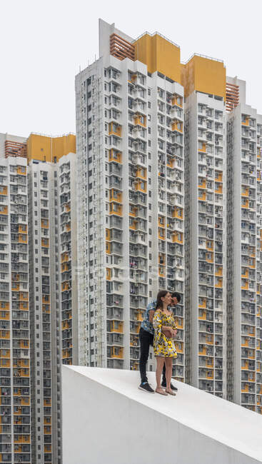 Cheerful adult man and faceless girl in colorful dress hugging together on concrete sloping roof against facades of spotted residential skyscrapers in Shek Kip Mei — Stock Photo
