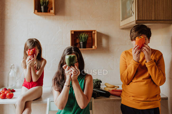 Mujer adulta y hermanos cubriendo la cara con pimientos de colores en la acogedora cocina en casa - foto de stock