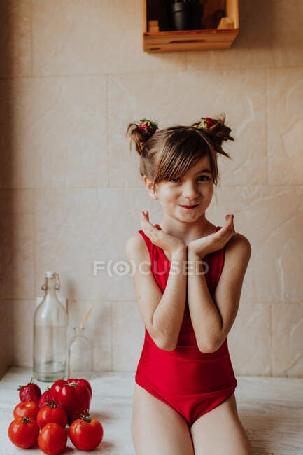Cute barefoot girl in red bodysuit sitting on counter with tomatoes and red pepper in kitchen — Stock Photo