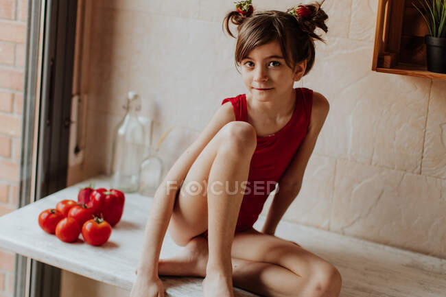 Jolie fille pieds nus en costume rouge assis sur le comptoir avec des tomates et du poivron rouge dans la cuisine — Photo de stock