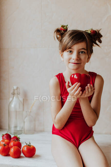 Menina descalça bonito em bodysuit vermelho e com morangos em seu cabelo segurando pimenta vermelha olhando para a câmera sentada no balcão perto de tomates na cozinha — Fotografia de Stock