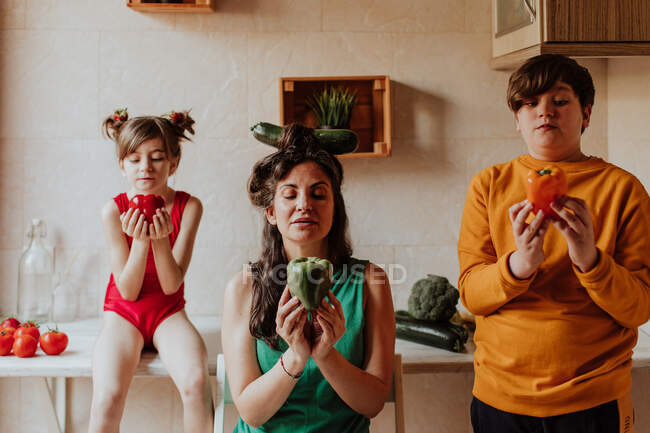Erwachsene Frau und Geschwister halten bunte Paprika in der Hand und beten zu Hause in der gemütlichen Küche — Stockfoto