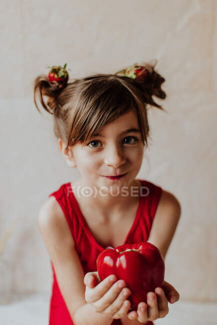 Adorable niña con fresas en el pelo mostrando pimienta fresca y mirando a la cámara - foto de stock