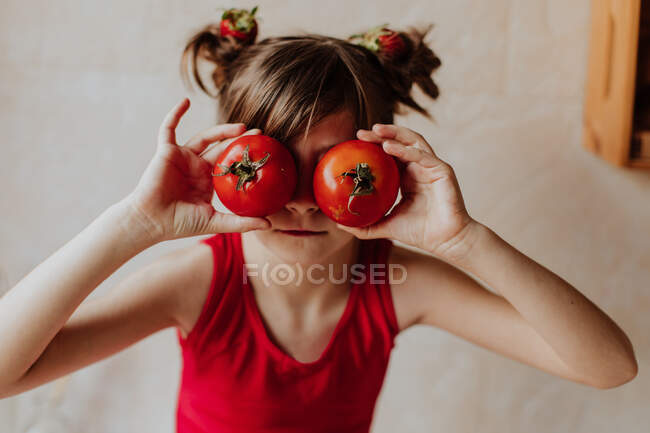 Nettes Mädchen hält frische Tomaten in der Nähe der Augen, während es in der Küche zu Hause Spaß hat — Stockfoto