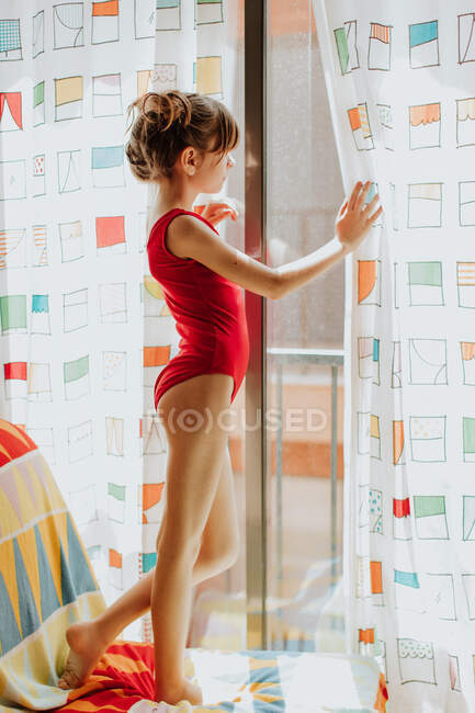 Side view of girl in red bodysuit standing on soft sofa and looking out window through colorful curtains at home — Stock Photo
