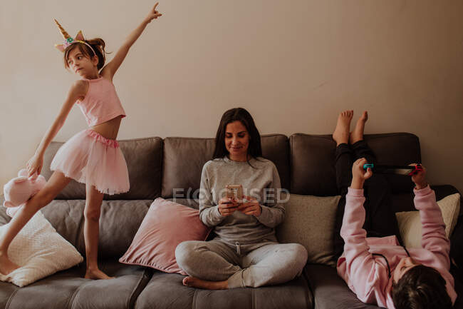 Mulher adulta que navega smartphone enquanto está sentada de pernas cruzadas no sofá perto da filha dançando e jogando filho em casa — Fotografia de Stock