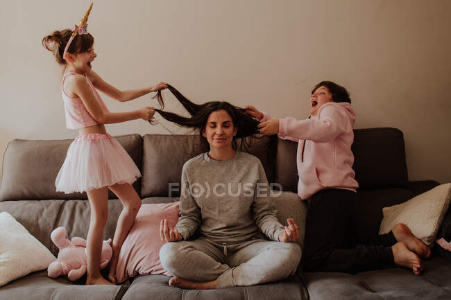 Excited little girl and teen boy pulling hair of adult woman sitting in Lotus pose on bed and meditating — Stock Photo