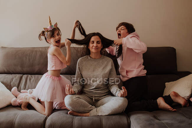 Excited little girl and teen boy pulling hair of adult woman sitting in Lotus pose on bed and meditating — Stock Photo